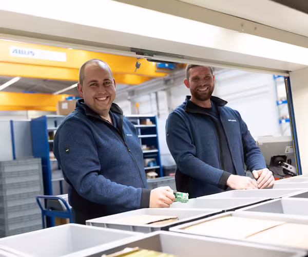 Zwei NEUERO Mitarbeiter stehen vor Ersatzteilkisten und lächeln in die Kamera.