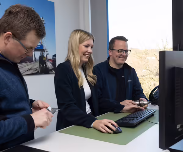 Drei NEUERO Mitarbeiter stehen lächelnd vor einem Bildschirm.
