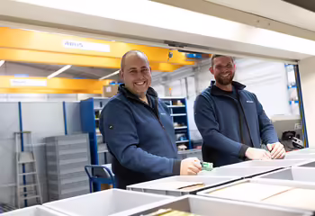 Zwei NEUERO Mitarbeiter stehen vor Ersatzteilkisten und lächeln in die Kamera.